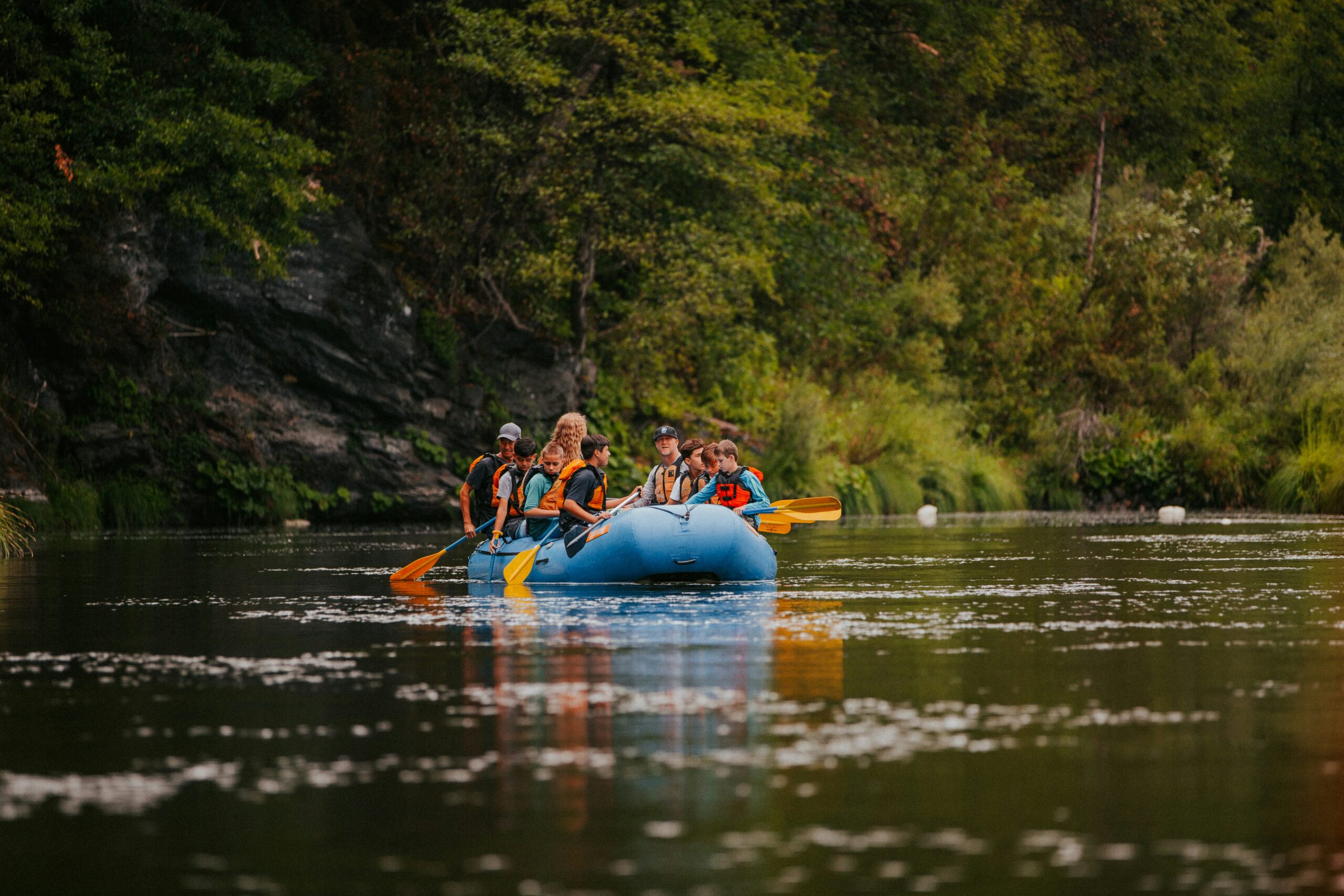 Image of a group of children and adults in orange lifejackets in a blue rafting boat along a tranquil river, trees lining the sides.