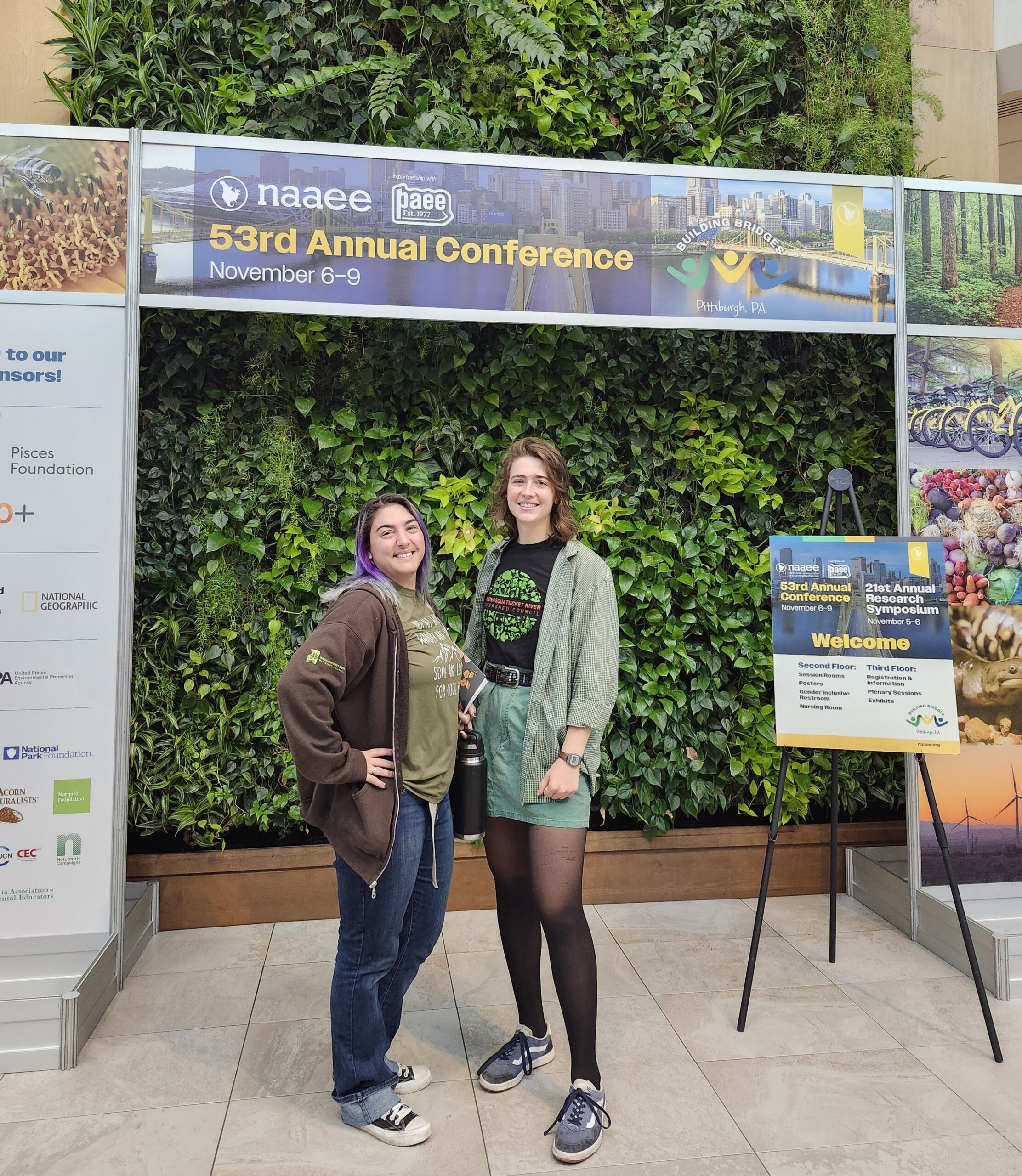 Amanda Peavey and Colleen Keenan standing in front of a green wall and a sign for the NAAEE Conference.