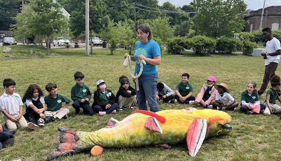 Educator standing outside with large model fish on ground, surrounded by group of children