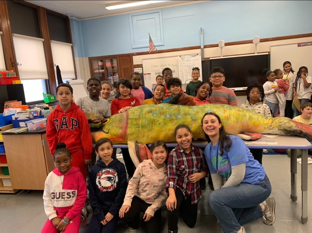 Students and teacher standing in classroom, surrounding a large colorful model of a fish