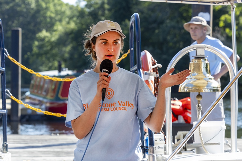 Person standing on a boat holding a microphone with river and captain in background